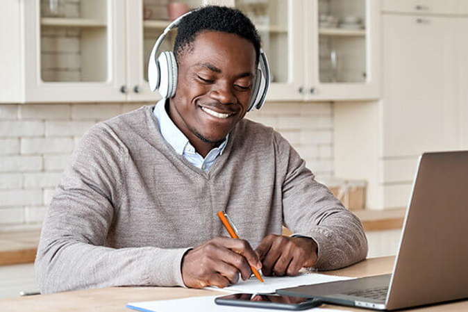 Young man wearing headphones smiling while taking notes in front of a laptop