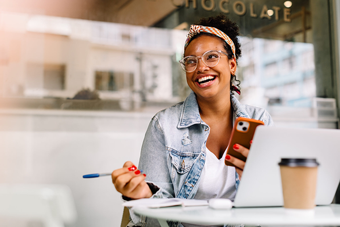 Young woman smiling outside a cafe with her phone while holding a pen in front of a laptop