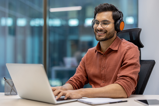 Young male entrepreneur using laptop and headphones at workplace, experiencing productive and focused work environment. Happy expression highlights satisfaction and positivity in professional setting