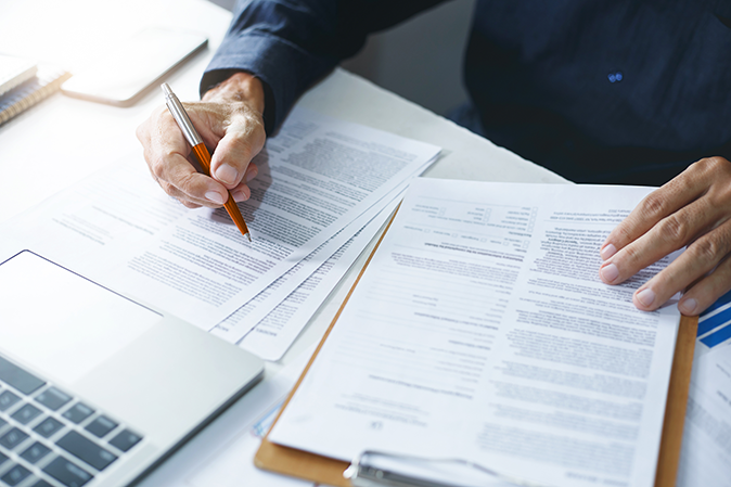 Person holding a pen while going over documents on a desk in front of a laptop