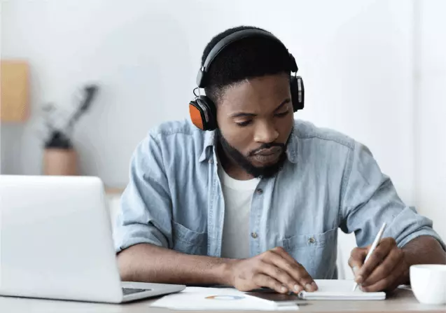 Man wearing headphones taking notes while studying at a desk with a laptop