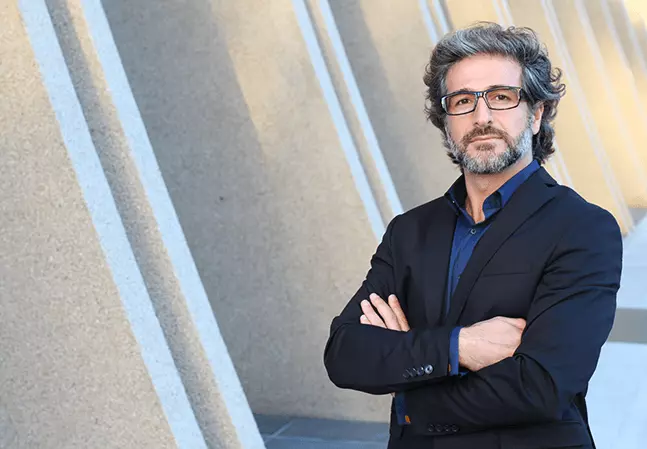 Bearded male law professor wearing glasses and a suit standing with arms crossed outside a building with concrete columns