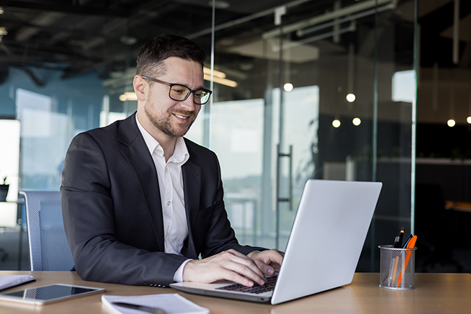 A smiling young man businessman in a business suit sits and works in the office on a laptop online, texts, chats with partners, clients.