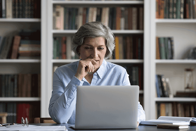 Senior woman sitting in front of laptop with bookshelves behind her