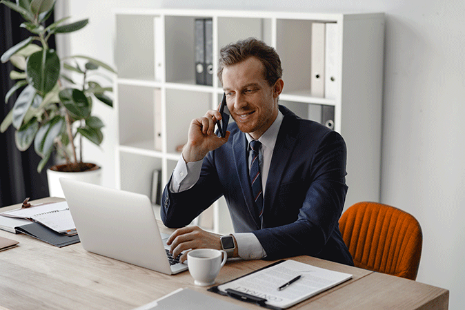 Middle man wearing a suit talking on the phone while typing on a laptop in his office