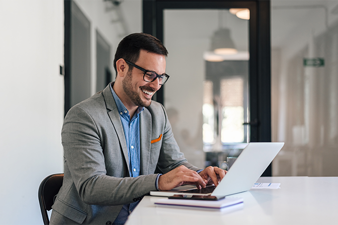 Cheerful young professional working on laptop. Smiling businessman is analyzing successful business plan. He is sitting at desk in corporate office.