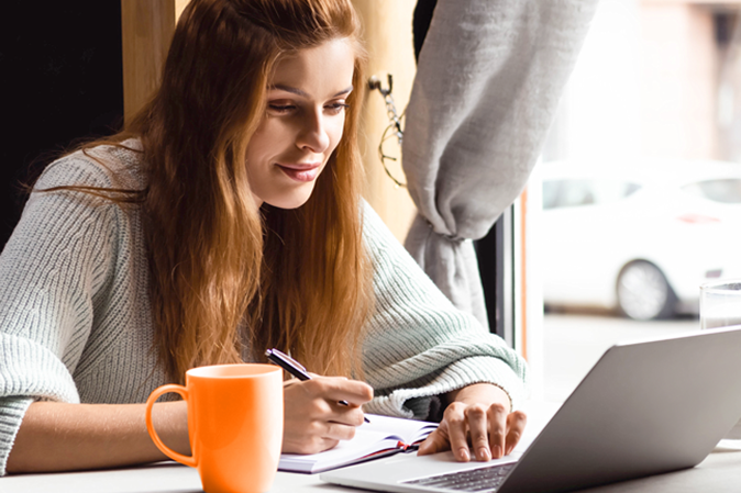 attractive woman writing in notepad while watching webinar on laptop in cafe