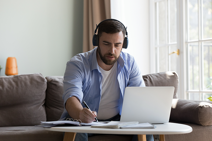 Young man at home wearing headphones working on a laptop and taking notes